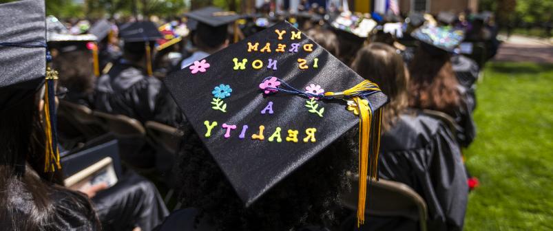 A student sits at their graduation, their cap reading, my dream is now a reality.